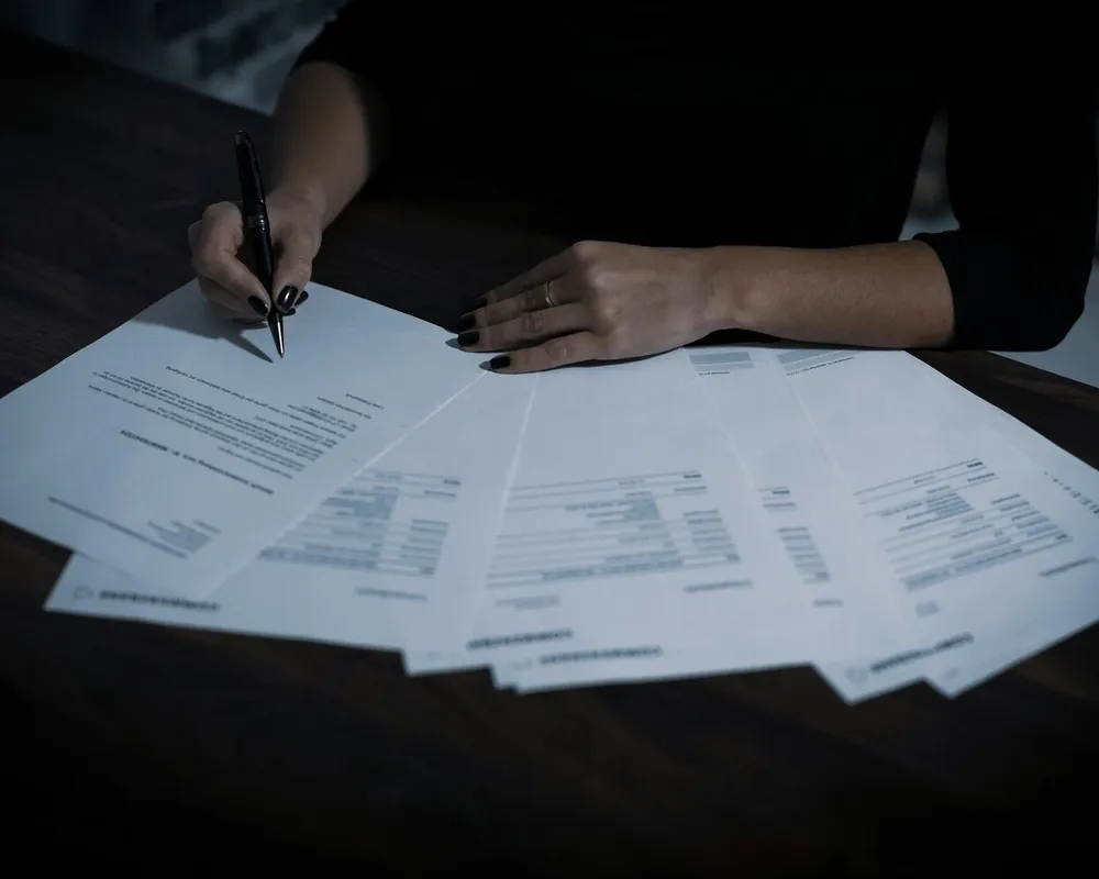 Un dossier de plusieurs feuillets étalé en éventail sur un bureau sombre, une main tenant un stylo au-dessus